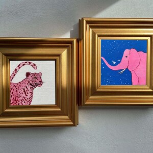 May include: Two framed art pieces. One features a pink leopard with a spotted pattern on a white background. The other depicts a pink elephant against a blue starry sky. Both are in gold frames, creating a cohesive art set.