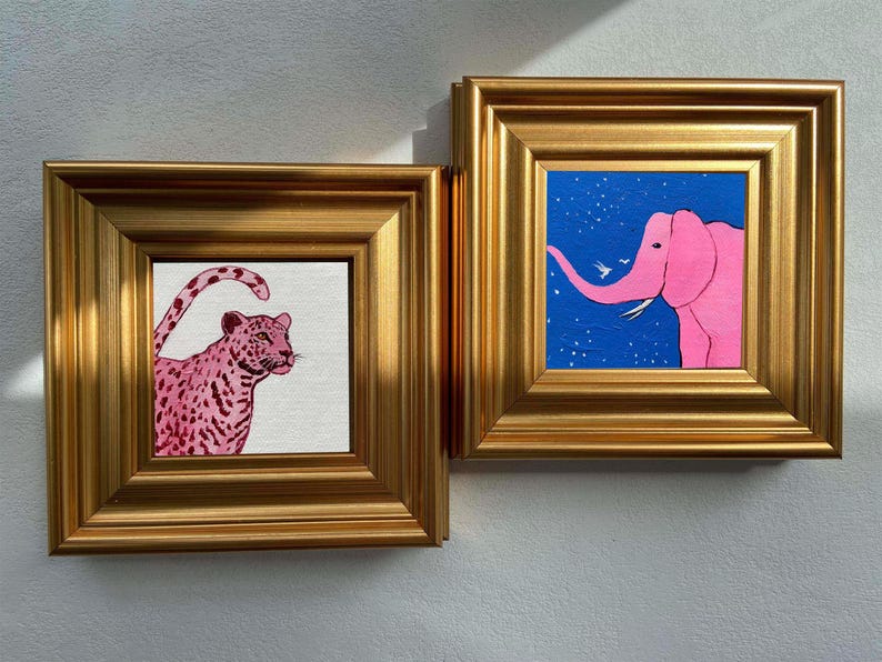 May include: Two framed art pieces. One features a pink leopard with a spotted pattern on a white background. The other depicts a pink elephant against a blue starry sky. Both are in gold frames, creating a cohesive art set.