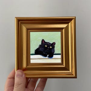 以下が含まれることがあります： 黄色い目を 가진黒猫の額入り絵画。猫は、明るい緑色の背景に対して、白い表面に休んでいます。フレームは金色で正方形です。
