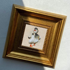 May include: A framed artwork featuring a whimsical illustration of a duckling. The duckling has gray and white feathers, orange feet, and a surprised expression. The artwork is set within a gold-colored, ornate frame.