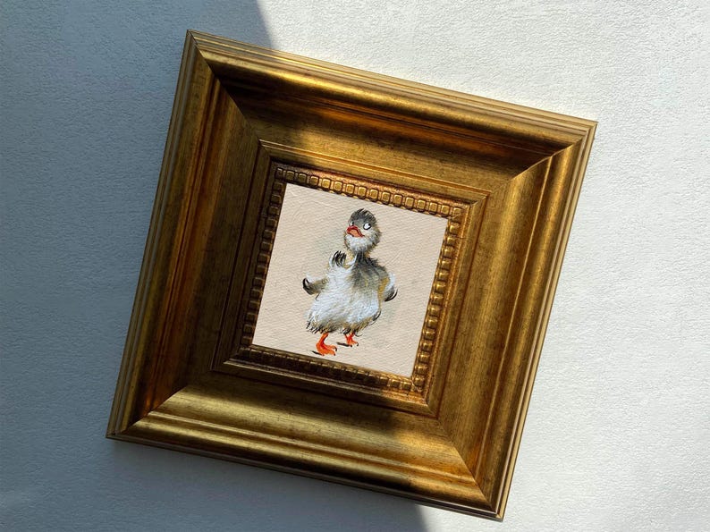 May include: A framed artwork featuring a whimsical illustration of a duckling. The duckling has gray and white feathers, orange feet, and a surprised expression. The artwork is set within a gold-colored, ornate frame.