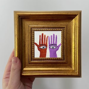 May include: A framed artwork featuring two hands, one red and one purple, each with an eye in the palm. The artwork is in a gold-colored frame. The hands are painted on a white background.