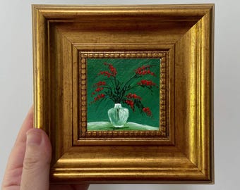 May include: A small framed painting depicting a still life of red flowers in a clear vase. The painting has a green background and a white tablecloth. The frame is gold-coloured and has a decorative edge.
