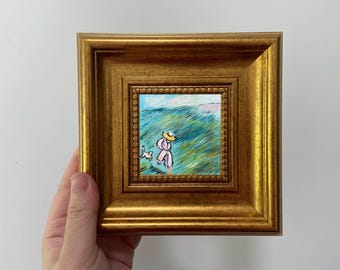 May include: A small framed painting with a gold-coloured ornate frame. The painting depicts a person in a hat and a dog walking through a field of green and blue brushstrokes. The artwork is held by a hand.