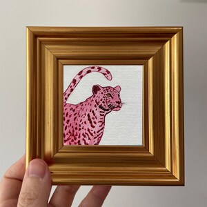 May include: A small, square painting of a pink leopard with dark pink spots, set in a gold-colored frame. The leopard is depicted in profile against a white background, with its tail curved upwards. The frame has a ridged design.