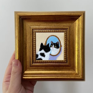 Puede incluir: Pintura enmarcada en oro que representa un gato blanco y negro mirándose en un espejo ovalado. El espejo tiene un fondo azul claro. Obra de arte con temática felina.