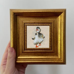 May include: A framed painting of a cartoon duckling with gray, white, and orange details. The duckling is centered on a beige background, framed by a gold, ornate frame. The painting is small, perfect for a desk or shelf.