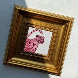 May include: A framed artwork featuring a pink leopard illustration. The leopard is depicted with a spotted pattern and is set against a white background. The frame is a gold color with a detailed, ornate design.