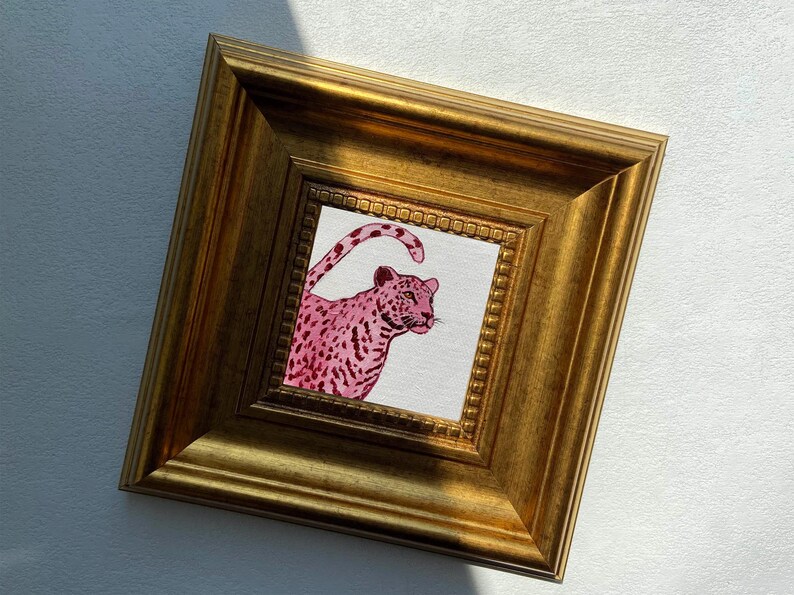 May include: A framed artwork featuring a pink leopard illustration. The leopard is depicted with a spotted pattern and is set against a white background. The frame is a gold color with a detailed, ornate design.