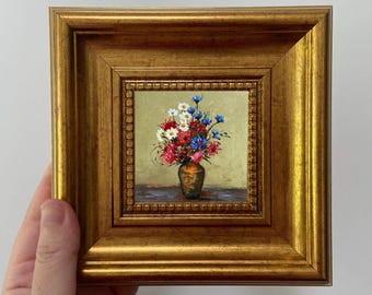 May include: A small, square painting in a gold frame. The artwork depicts a vase of colorful flowers, including red, white, and blue blooms. The vase is a dark bronze color, and the background is a soft yellow.