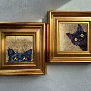May include: Two framed cat portraits. Each features a black cat with yellow eyes peeking out from a gold-colored background. The frames are ornate, gold-colored, and square. The artwork is mounted on a white wall.