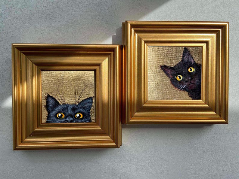 May include: Two framed cat portraits. Each features a black cat with yellow eyes peeking out from a gold-colored background. The frames are ornate, gold-colored, and square. The artwork is mounted on a white wall.