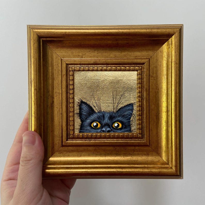 May include: A small, square painting of a black cat's face peeking over a gold background. The cat has bright yellow eyes and is framed in a gold, ornate wooden frame. The artwork is held by a hand.