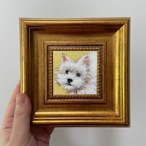 May include: A small, framed painting of a white dog with a fluffy coat and dark eyes. The artwork is set against a yellow background and encased in a gold-colored, ornate frame. The frame is rectangular and has a detailed, textured design.