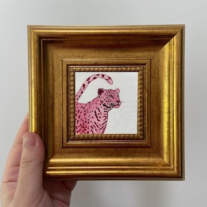 May include: A small square painting of a pink leopard with dark pink spots, set in a gold-colored ornate frame. The leopard is depicted on a white background, with its tail curved upwards. The frame has a textured, detailed design.