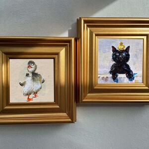 May include: Two framed paintings with gold frames. One features a duck with gray and white feathers, orange feet, and a red beak. The other depicts a black cat wearing a yellow crown. Both paintings are on a white wall.