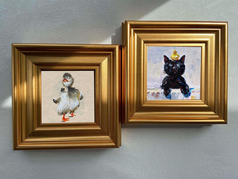 May include: Two framed paintings with gold frames. One features a duck with gray and white feathers, orange feet, and a red beak. The other depicts a black cat wearing a yellow crown. Both paintings are on a white wall.