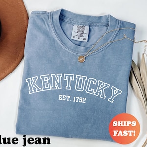 Może przedstawiać: Niebieska koszulka dżinsowa z napisem "KENTUCKY" białymi literami i "EST. 1792" poniżej. Złoty naszyjnik z wisiorkiem spoczywa na koszulce. Pomarańczowa naklejka z napisem "SHIPS FAST!" znajduje się po prawej stronie.