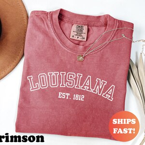 May include: Crimson-colored t-shirt with the word "LOUISIANA" in white block letters and "EST. 1812" underneath. The shirt has a crew neck and a small tag. A gold necklace with a pendant is draped across the shirt. The shirt is labeled "SHIPS FAST!"