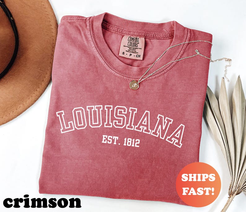 May include: Crimson-colored t-shirt with the word "LOUISIANA" in white block letters and "EST. 1812" underneath. The shirt has a crew neck and a small tag. A gold necklace with a pendant is draped across the shirt. The shirt is labeled "SHIPS FAST!"