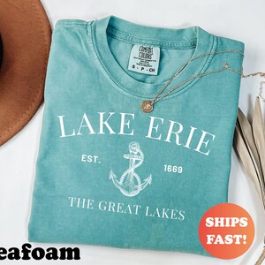 May include: Seafoam green t-shirt with the text "LAKE ERIE" and an anchor graphic. The shirt also includes the text "EST. 1669" and "THE GREAT LAKES". A gold necklace with a pendant is on the shirt.