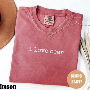 May include: Crimson-colored t-shirt with the phrase "i love beer" in white lettering. The shirt has a crew neck and a small tag at the neckline. A gold necklace with a pendant is draped on the shirt. A brown hat is in the background.
