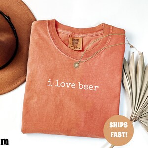 May include: Coral-colored t-shirt with the phrase "i love beer" in white lettering. A gold necklace with a pendant rests on the shirt. A brown cowboy hat and dried palm leaves are also in the image.