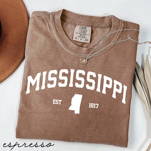 May include: Espresso-colored t-shirt with the word "MISSISSIPPI" in white block letters. Below the word is "EST" and "1817" with a white outline of the state. A silver necklace with a pendant rests on the shirt.
