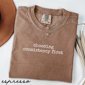 Puede incluir: Camiseta color espresso con la frase "choosing consistency first" en blanco. Un collar plateado con un pequeño colgante descansa sobre la camiseta. La camiseta es de la marca Comfort Colors, talla S-P-CH.