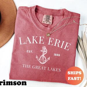 May include: Crimson-colored t-shirt with the text "LAKE ERIE" and an anchor graphic. The shirt also includes the text "EST. 1669" and "THE GREAT LAKES". A gold necklace is draped on the shirt. The shirt has a round neck.