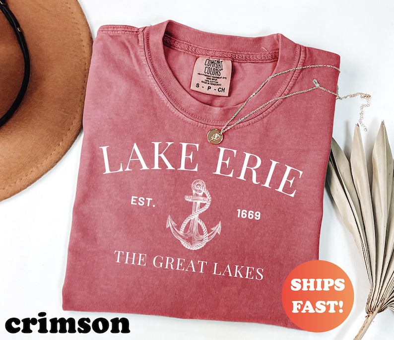 May include: Crimson-colored t-shirt with the text "LAKE ERIE" and an anchor graphic. The shirt also includes the text "EST. 1669" and "THE GREAT LAKES". A gold necklace is draped on the shirt. The shirt has a round neck.