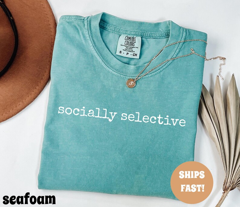 May include: Seafoam green t-shirt with the words "socially selective" printed in white. A gold necklace with a pendant rests on the shirt. The shirt is folded and has a Comfort Colors tag. A brown hat is visible in the background.