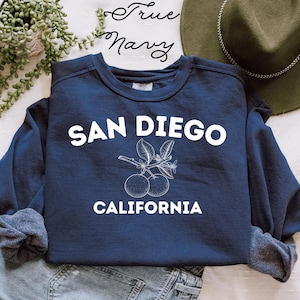 May include: Navy blue sweatshirt featuring the text "SAN DIEGO CALIFORNIA" in white. A white illustration of oranges and leaves is between the city and state names. The words "True Navy" are written in cursive at the top of the image.
