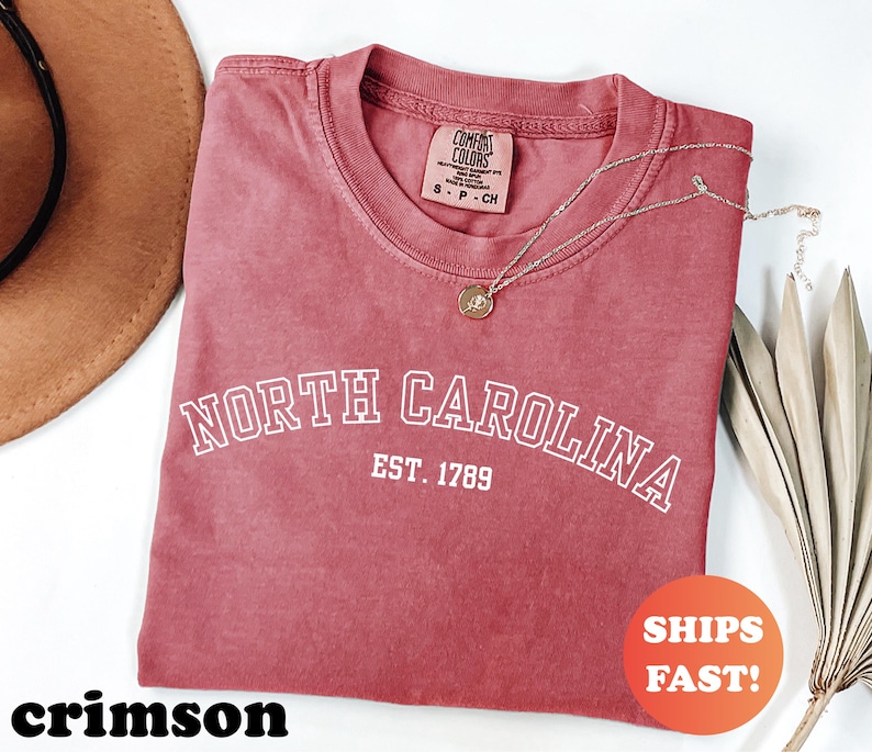 Puede incluir: Camiseta de color carmes&iacute; con las palabras "NORTH CAROLINA" y "EST. 1789" impresas en blanco. La camiseta tiene cuello redondo y una peque&ntilde;a etiqueta en el cuello. Un collar dorado con un colgante est&aacute; drapeado sobre la camiseta. Las palabras "SHIPS FAST!" est&aacute;n en un c&iacute;rculo naranja.