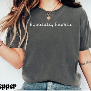 Może przedstawiać: Ciemnoszara koszulka z białym napisem "Honolulu, Hawaii". Koszulka ma okrągły dekolt i wydaje się być wykonana z miękkiego materiału. Słowo "pepper" znajduje się na dole obrazu.