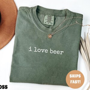May include: A moss green t-shirt with the phrase "i love beer" in white lettering. A gold necklace with a pendant rests on the shirt. The shirt is folded and has a tag on the collar.