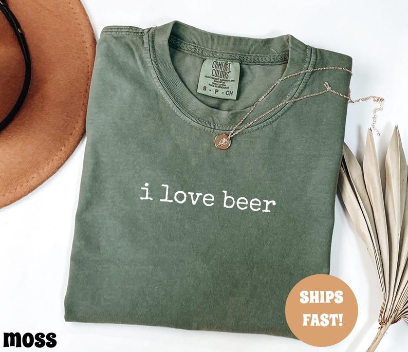 May include: A moss green t-shirt with the phrase "i love beer" in white lettering. A gold necklace with a pendant rests on the shirt. The shirt is folded and has a tag on the collar.