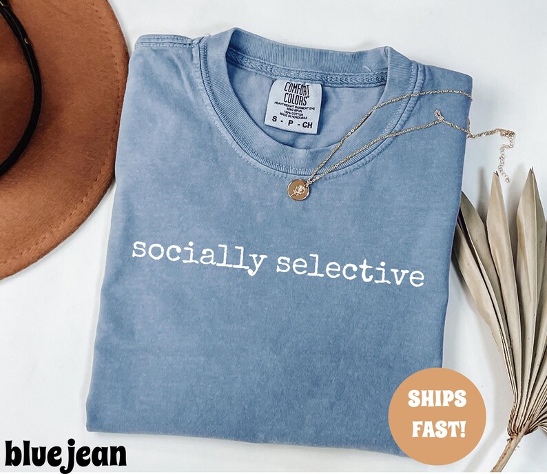 May include: A blue jean Comfort Colors t-shirt with the words "socially selective" printed in white. A gold necklace with a pendant rests on the shirt. A brown hat and dried leaves are in the background.