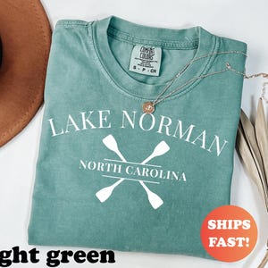 May include: A light green t-shirt with the text "LAKE NORMAN NORTH CAROLINA" and crossed oars in white. A gold necklace rests on the shirt. The shirt has a Comfort Colors tag. An orange sticker with "SHIPS FAST!" is visible.
