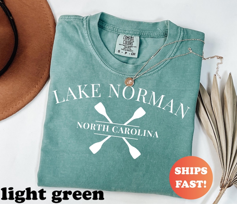 May include: A light green t-shirt with the text "LAKE NORMAN NORTH CAROLINA" and crossed oars in white. A gold necklace rests on the shirt. The shirt has a Comfort Colors tag. An orange sticker with "SHIPS FAST!" is visible.