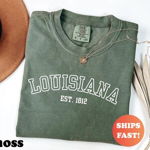May include: A moss green t-shirt with the word "LOUISIANA" in white block letters and "EST. 1812" underneath. A gold necklace with a pendant rests on the shirt. An orange circle with the words "SHIPS FAST!" is in the lower right corner.