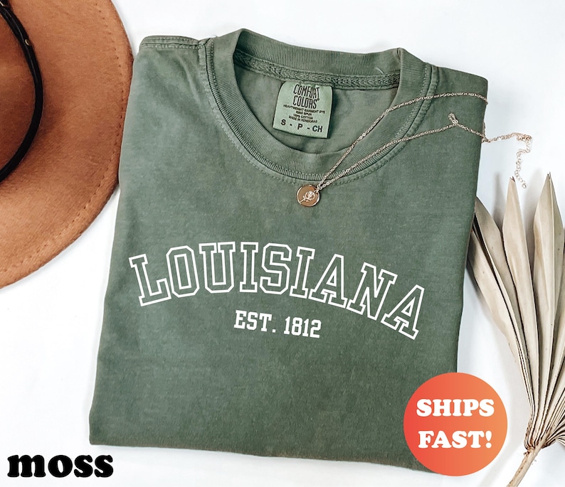 May include: A moss green t-shirt with the word "LOUISIANA" in white block letters and "EST. 1812" underneath. A gold necklace with a pendant rests on the shirt. An orange circle with the words "SHIPS FAST!" is in the lower right corner.