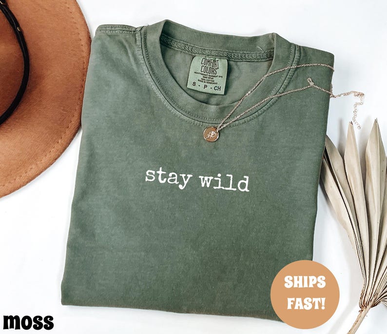 May include: A moss green t-shirt with the words "stay wild" in white lettering. A gold necklace with a pendant rests on the shirt. The shirt has a crew neck and a tag that reads "Comfort Colors".