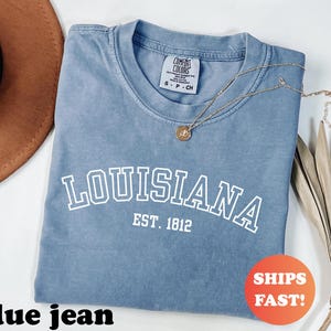 May include: A blue jean colored t-shirt with the word "LOUISIANA" printed in white, arched across the front, with "EST. 1812" below. A gold necklace with a pendant rests on the shirt. The shirt has a Comfort Colors tag.