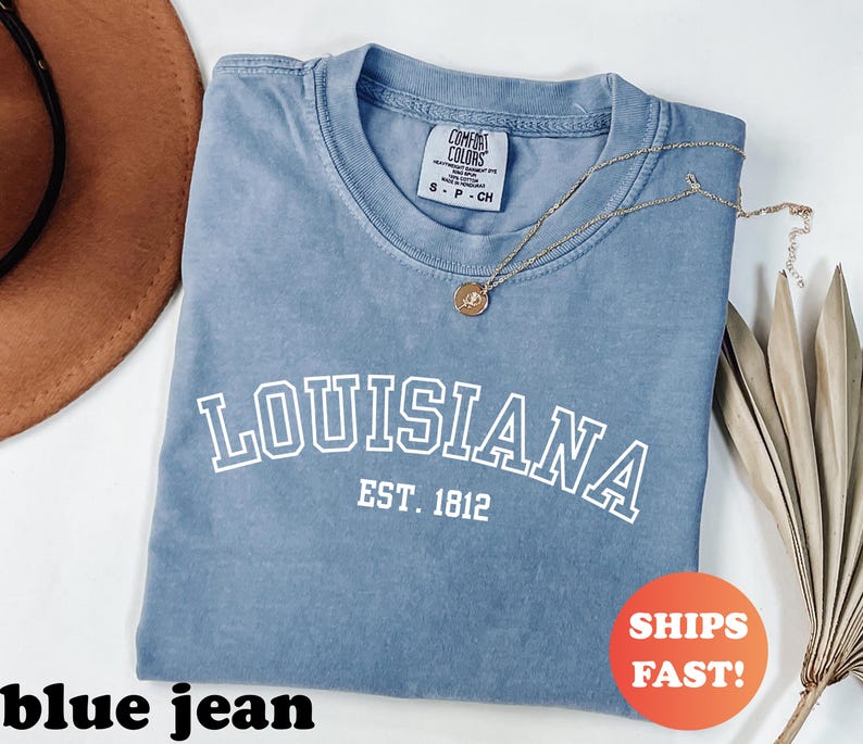 May include: A blue jean colored t-shirt with the word "LOUISIANA" printed in white, arched across the front, with "EST. 1812" below. A gold necklace with a pendant rests on the shirt. The shirt has a Comfort Colors tag.