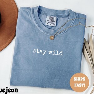May include: A blue jean colored t-shirt with the words "stay wild" in white lettering. A gold necklace with a circular pendant rests on the shirt. The shirt is folded and placed on a white surface.