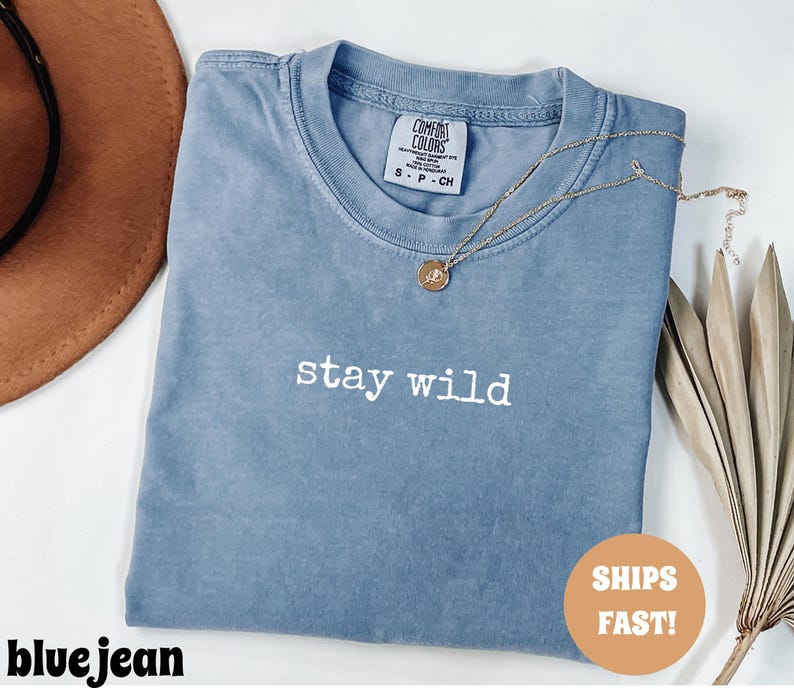 May include: A blue jean colored t-shirt with the words "stay wild" in white lettering. A gold necklace with a circular pendant rests on the shirt. The shirt is folded and placed on a white surface.
