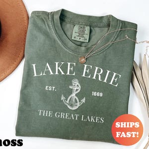 May include: A moss green t-shirt with the text "LAKE ERIE" and an anchor graphic. The shirt also features the text "EST. 1669" and "THE GREAT LAKES". A gold necklace with a pendant is on the shirt. An orange circle with "SHIPS FAST!" is in the image.