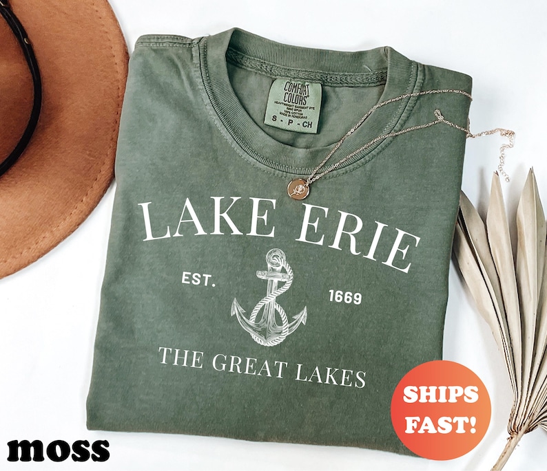 May include: A moss green t-shirt with the text "LAKE ERIE" and an anchor graphic. The shirt also features the text "EST. 1669" and "THE GREAT LAKES". A gold necklace with a pendant is on the shirt. An orange circle with "SHIPS FAST!" is in the image.