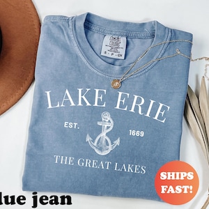May include: A blue jean colored t-shirt with the words "LAKE ERIE" and an anchor graphic. The text "EST. 1669 THE GREAT LAKES" is also printed on the shirt. A gold necklace and a brown hat are also visible.